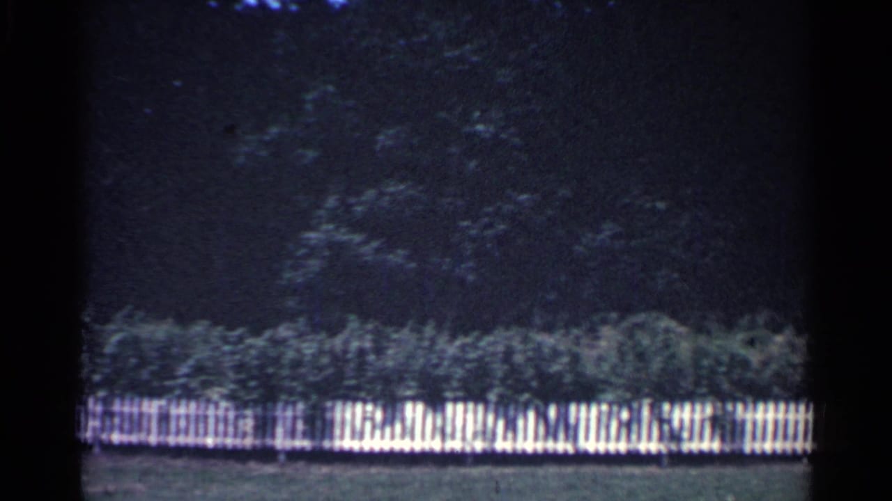 1959 Ohio home-movie frame with a long white picket fence, low greenery, and dense trees behind the yard.