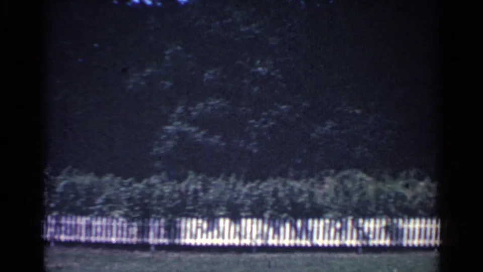 1959 Ohio home-movie frame with a long white picket fence, low greenery, and dense trees behind the yard.