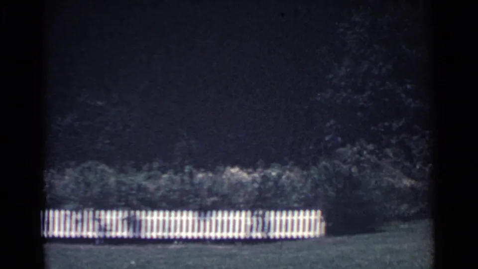 1959 Ohio home-movie frame showing the end of a white picket fence beside shrubs, lawn, and dense trees.