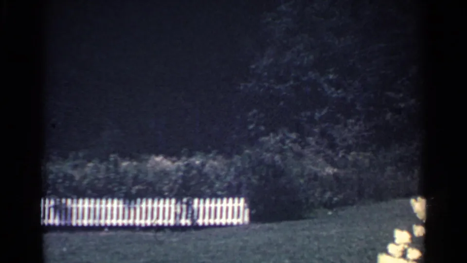 1959 Ohio home-movie frame showing a white picket fence across a shaded backyard lawn with trees behind it.