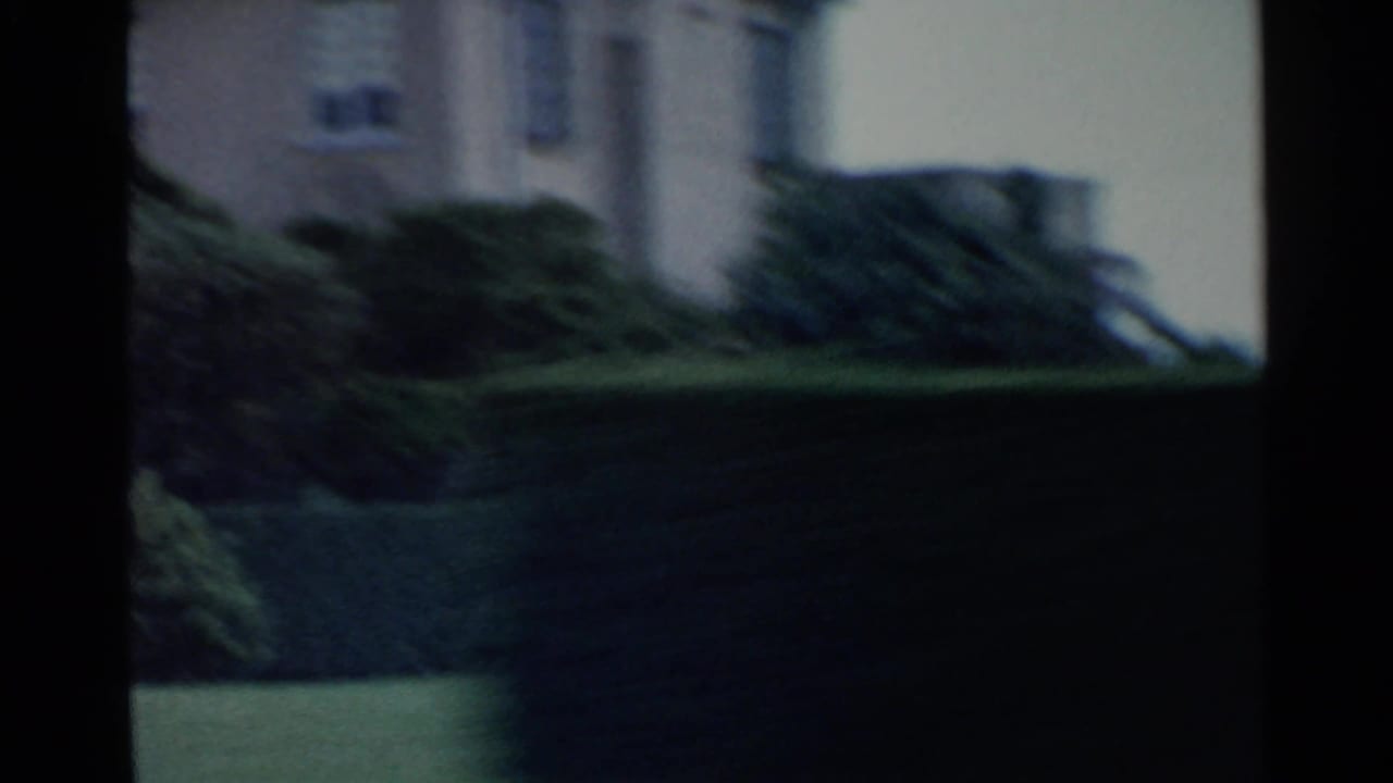 Soft, underexposed home-movie frame showing greenery and a residential wall during a quick handheld move.
