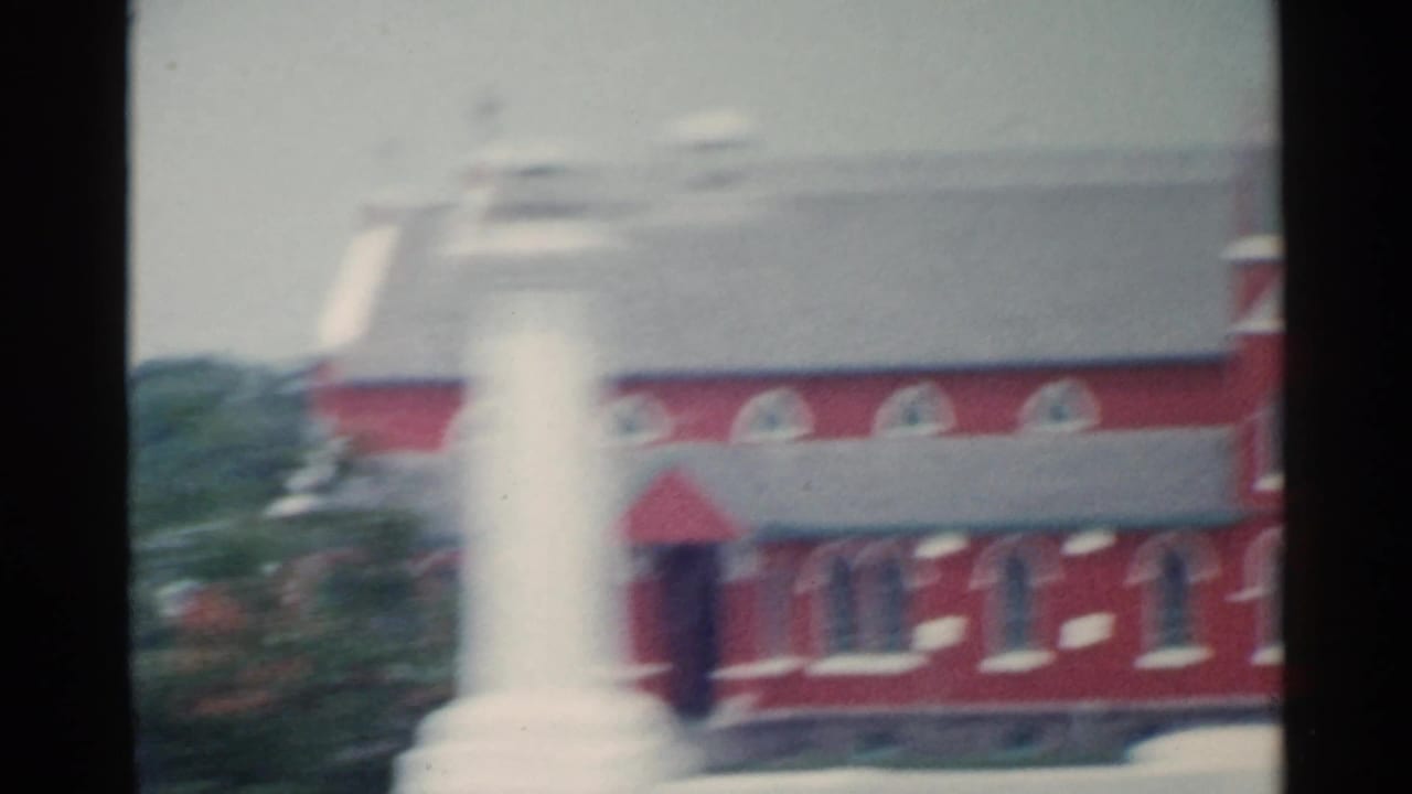 Moving-car view of a red community or church-like building with arched windows and a white monument in front, recorded in 1959 San Francisco.