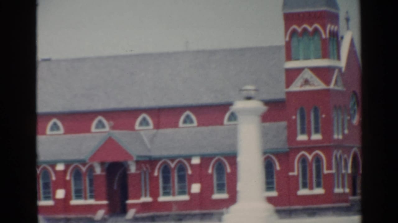 Clearer 1959 San Francisco frame of a red church-like building with arched windows, gray rooflines, and a white monument near the foreground.