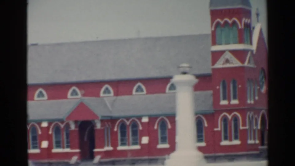 Clearer 1959 San Francisco frame of a red church-like building with arched windows, gray rooflines, and a white monument near the foreground.