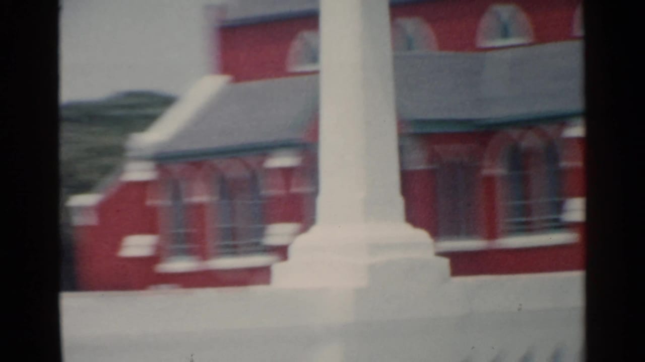 White monument or cross in front of a red brick church wall and windows.