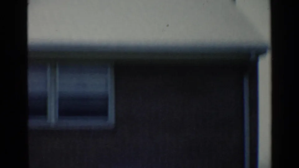 Soft, shaky 1959 San Francisco-area frame showing part of a low building, a small window, roof edge, and dark vertical film borders.