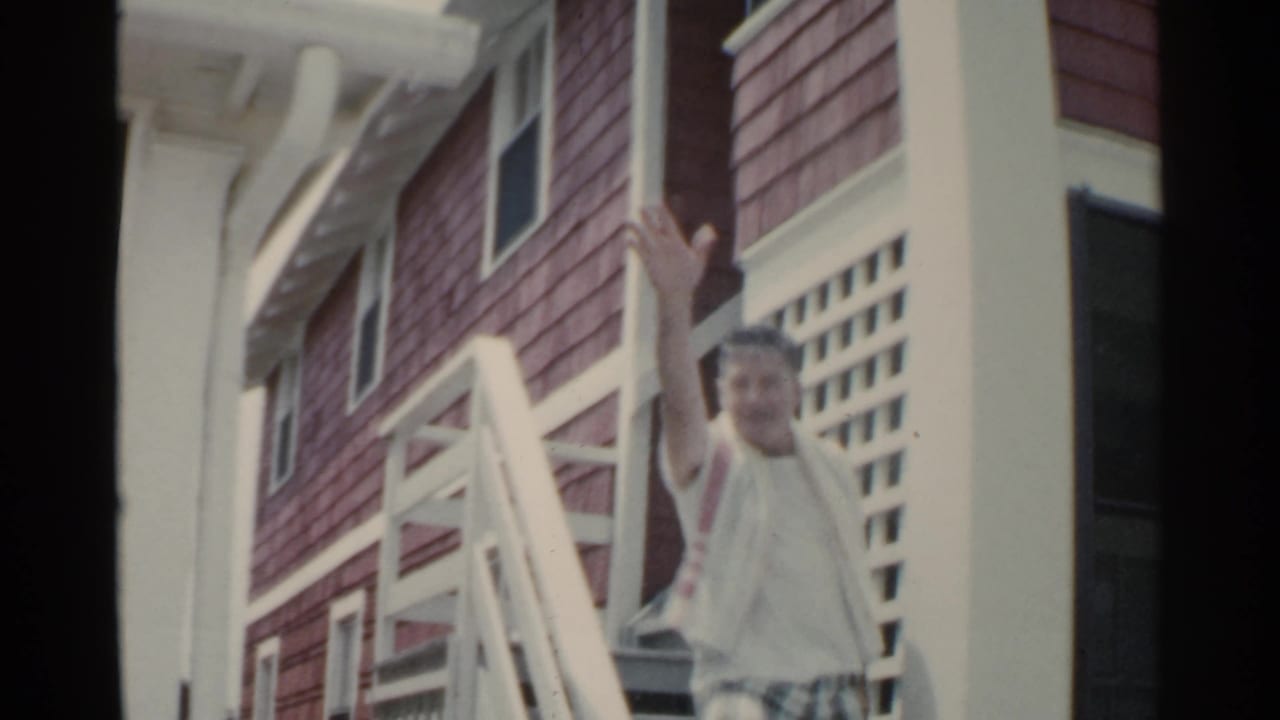 Man in summer clothing waves from a porch beside a red-shingled building.
