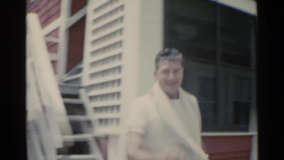A smiling man with a towel over his shoulder stands by white porch railings in the 1959 San Francisco-labeled vacation footage.