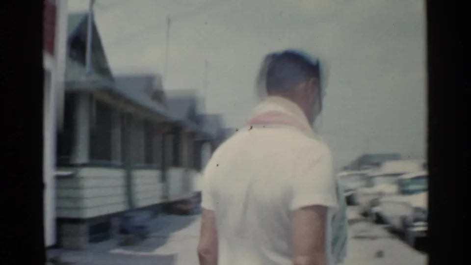 The man is seen from behind walking away along a residential street, with houses and parked cars visible in the soft 8mm frame.