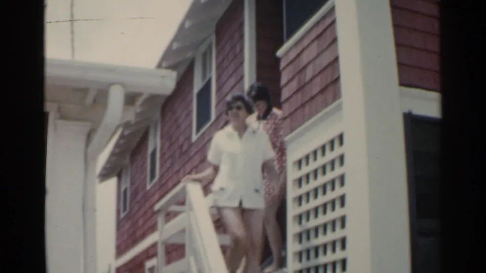 Two adults move down the white porch steps outside the red-shingled building in the 1959 San Francisco-labeled home movie.