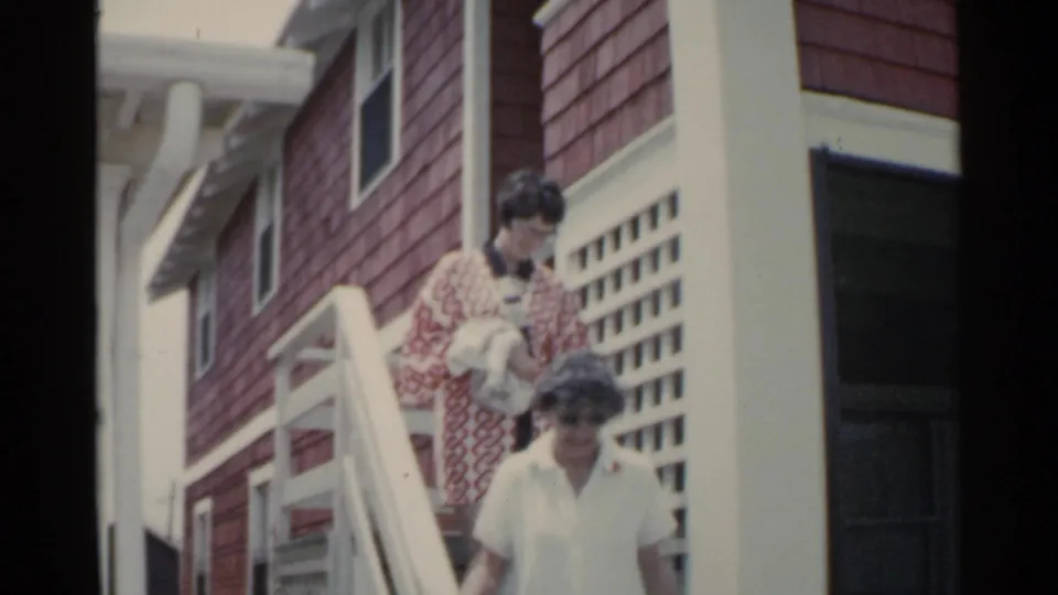 Two women descend the white railing staircase outside a red-shingled San Francisco house, one carrying a light towel or garment.