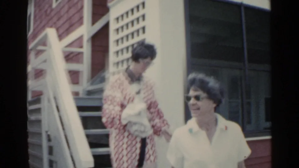 A woman in a white short-sleeved top and sunglasses passes near the camera while another woman in a red patterned robe holds a towel by the stairs.