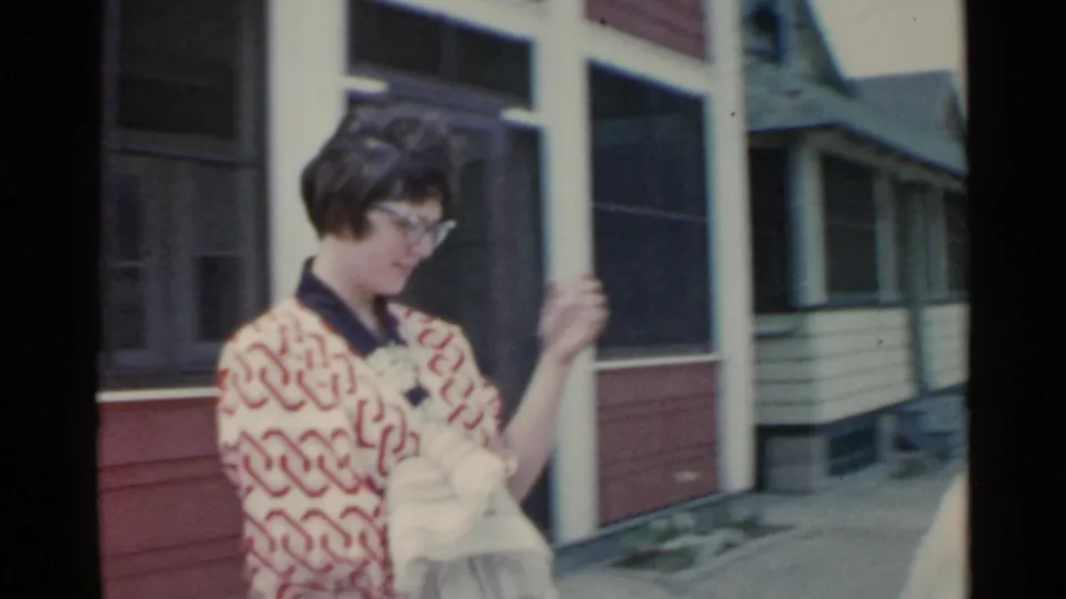 A woman in a red and white patterned robe looks down at her hands while holding a towel near screened porches and neighboring house fronts.
