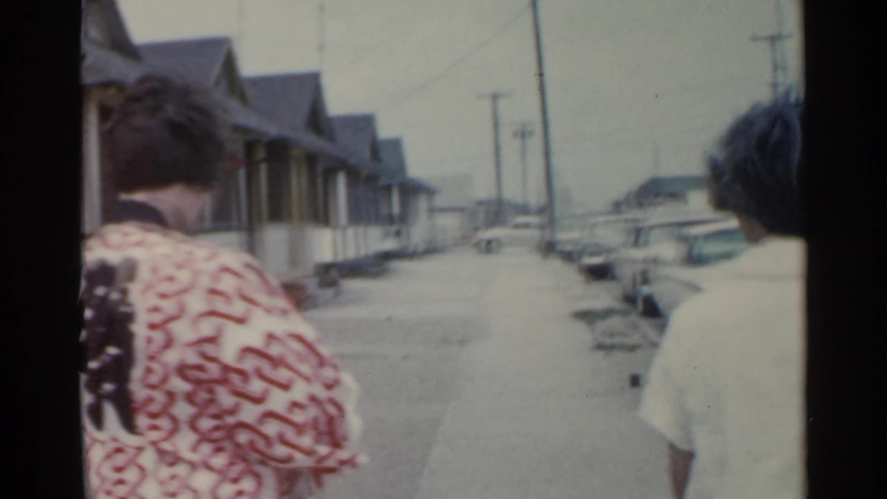 Two women seen from behind walking along a residential sidewalk with houses and parked cars.