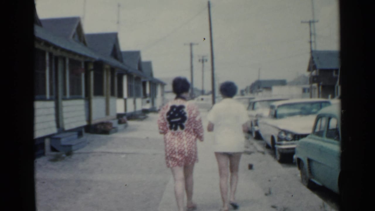 Two women are filmed from behind on a residential San Francisco sidewalk in 1959, with older cars and repeated house fronts visible.