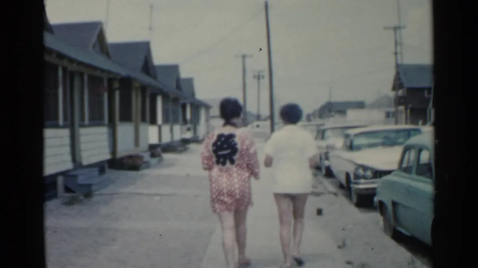 Two women are filmed from behind on a residential San Francisco sidewalk in 1959, with older cars and repeated house fronts visible.