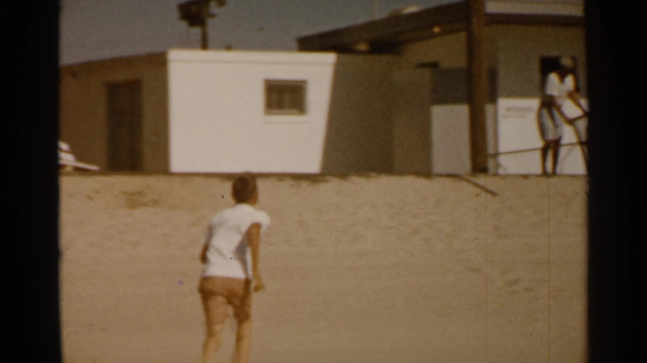 A later 8mm frame shows a person moving uphill toward a plain building, with two distant figures near exterior stairs.