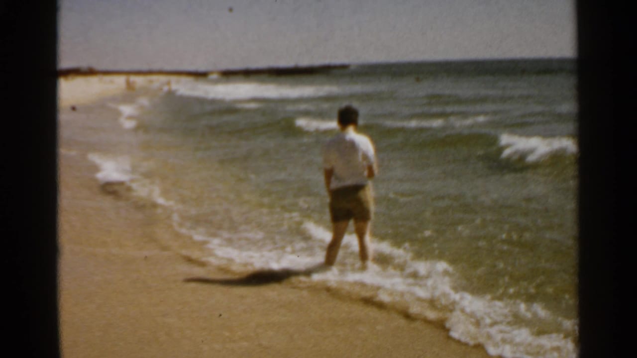 Person in a white shirt stands at the edge of the surf on a sandy beach.