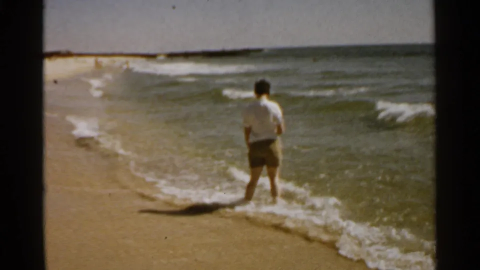1959 San Francisco beach footage shows a person in a white shirt and shorts standing where small waves meet the sand.