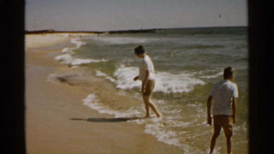 Two people in light shirts and shorts are near the San Francisco shoreline as surf washes onto the sand.