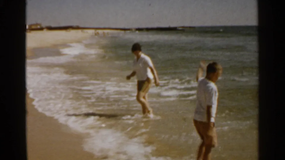 Two beachgoers in 1959 San Francisco step near foamy surf, with the ocean and sandy shore filling the frame.