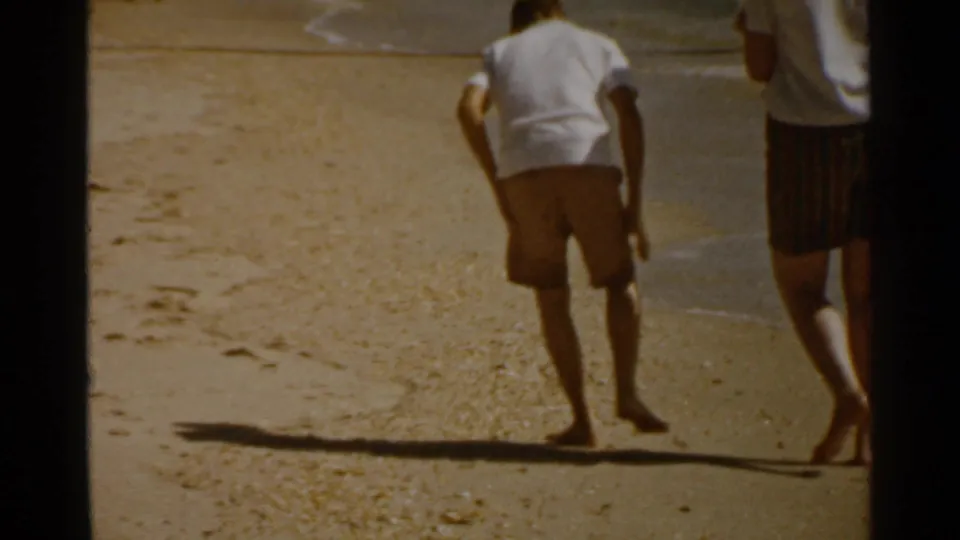 A handheld 1959 San Francisco beach frame shows partly cropped figures walking on sand beside the waterline.