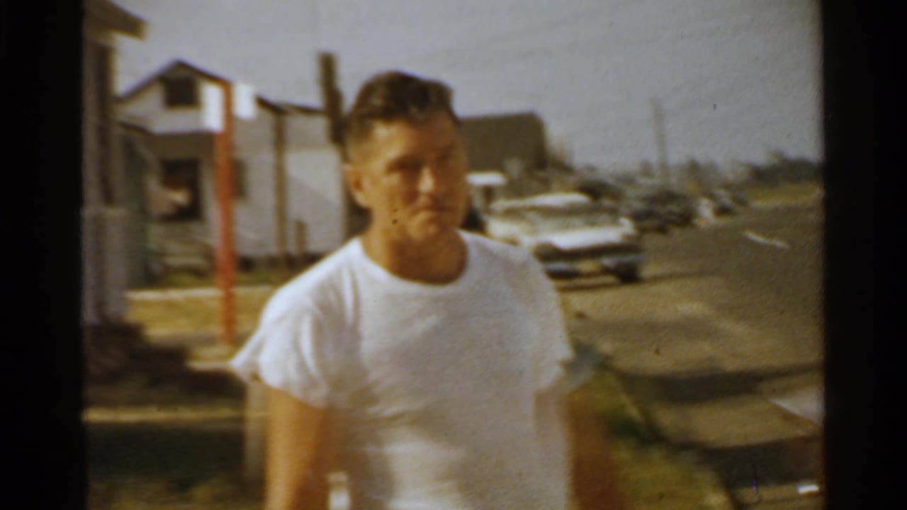 A man in a white T-shirt walks on a San Francisco residential street in 1959, with parked cars and houses behind him.