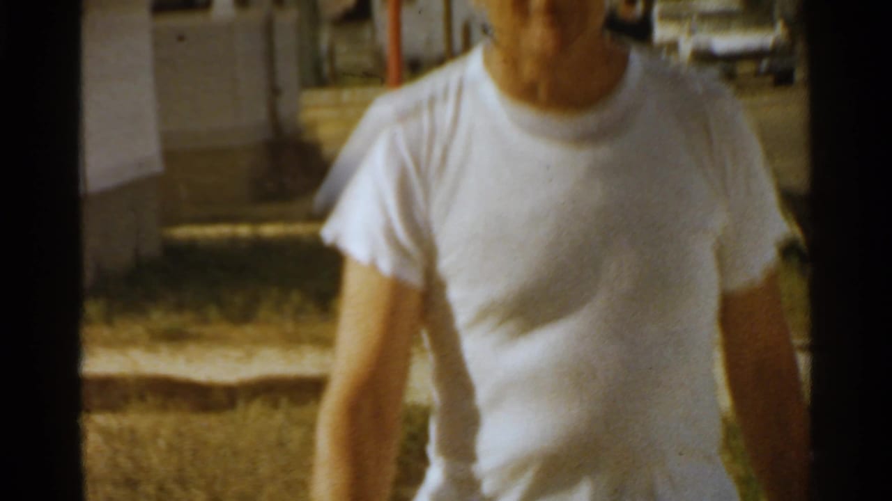 Close 1959 San Francisco street view of a man in a white T-shirt walking past a modest house and patch of grass.