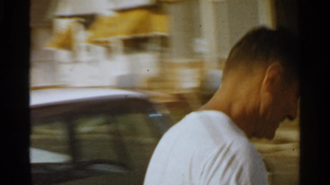 Man in a white T-shirt beside a parked car on a blurred street.