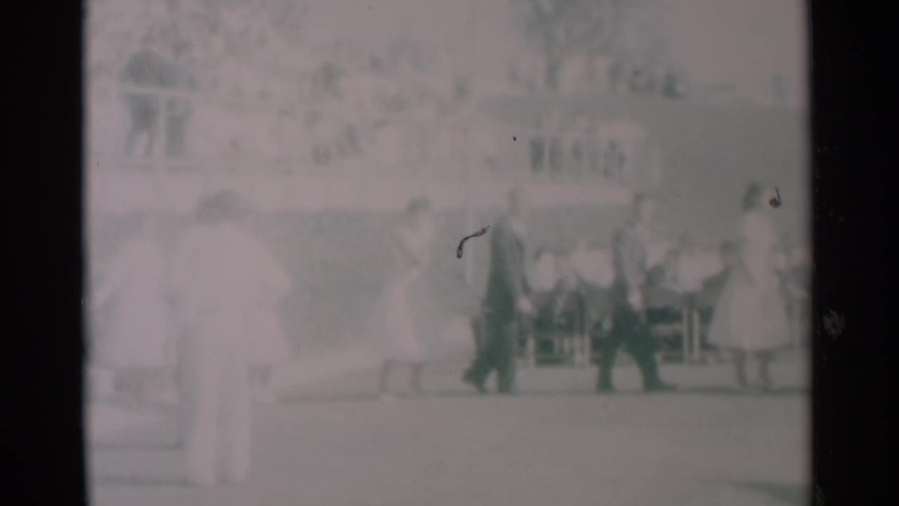 People in dresses and suits walk past seated onlookers at an outdoor gathering.