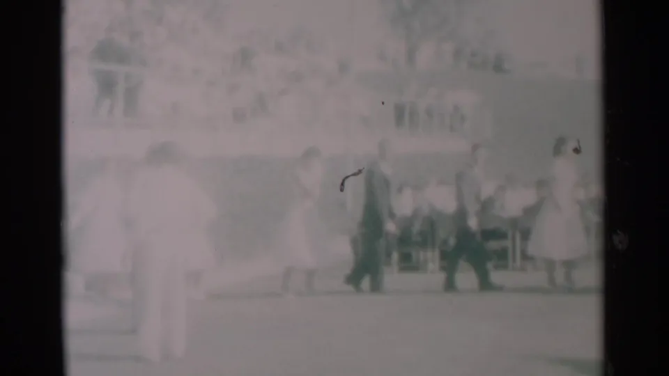 1959 South Dakota 8mm frame showing people in light dresses and dark suits walking past tables with seated onlookers.
