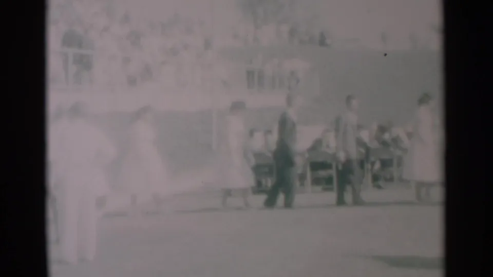 1959 South Dakota home-movie frame with a small group walking in single file past seated people at outdoor tables.