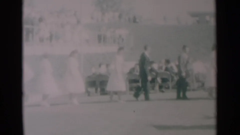 Soft 8mm frame from South Dakota in 1959 showing walkers in dresses and suits crossing before seated onlookers.