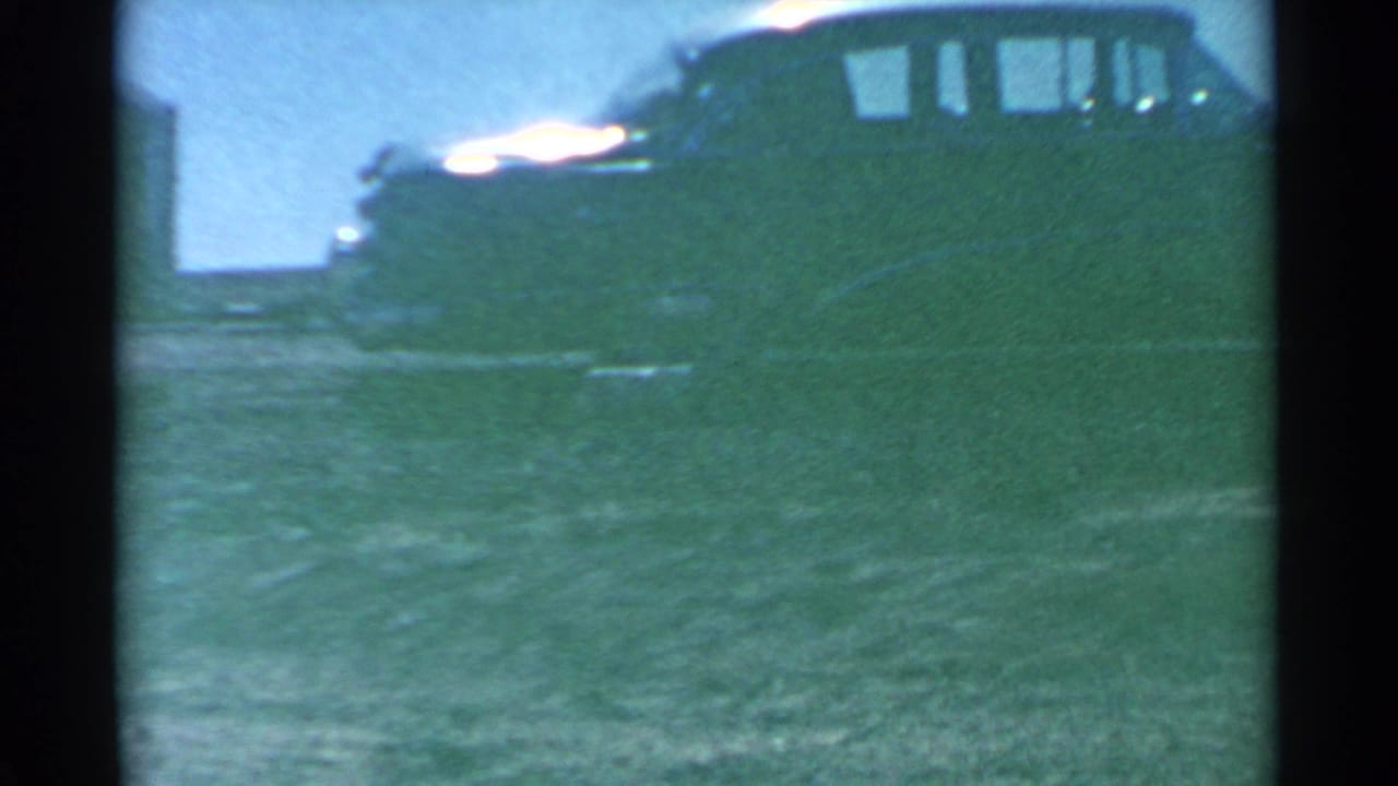 Parked dark vehicle beside open grass, seen through heavy 8mm grain.