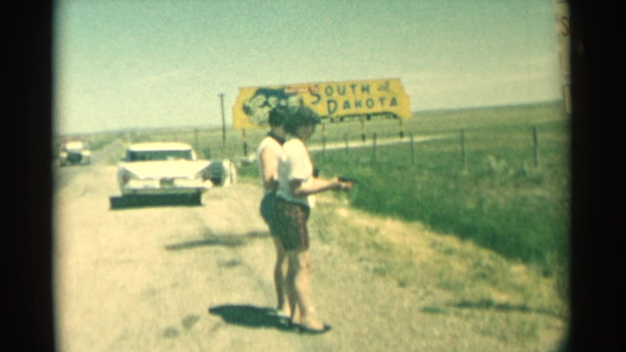 Two women stand by a rural road near a white car and South Dakota sign.