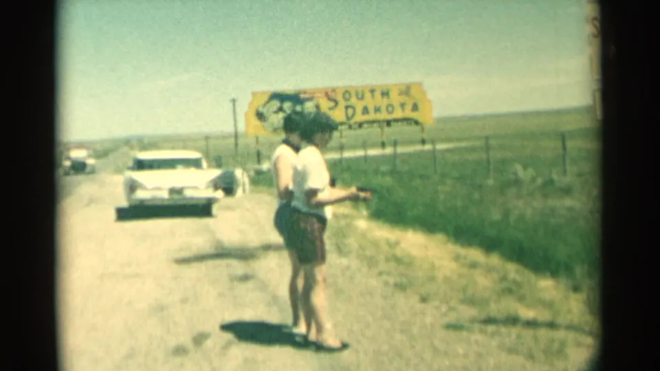 Two women on a rural South Dakota roadside in 1959, with one holding a handgun near a white car and yellow state sign.