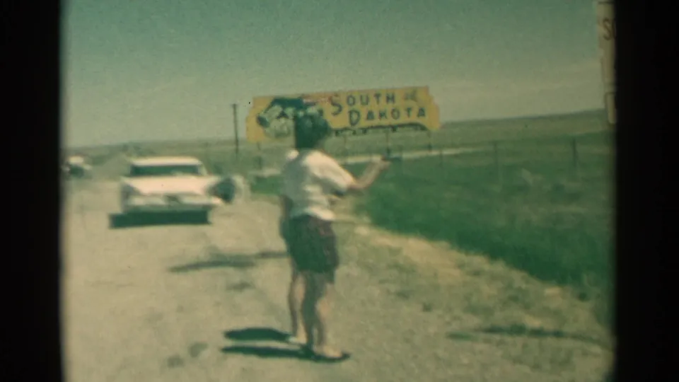 A woman points a black handgun on a rural South Dakota roadside, with another woman nearby and a 1950s white car behind them.