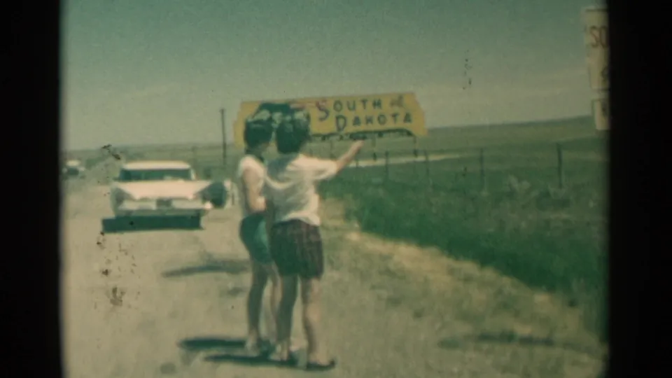 Two women stand on a rural South Dakota road shoulder in 1959 while one gestures with a handgun near the yellow state sign.