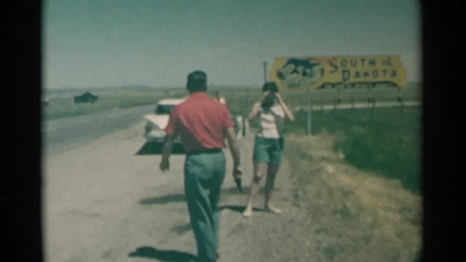 1959 home-movie frame of two adults stopping near a yellow South Dakota border sign on a rural roadside with a white car nearby.