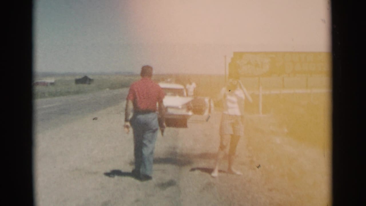 A man walks toward a white car while a woman stands near the South Dakota sign.