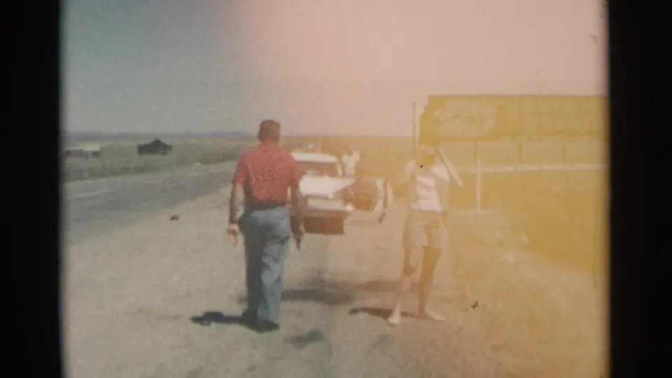 Faded 8mm frame of a roadside stop in South Dakota, with a man walking toward a white car and a woman near the yellow state sign.