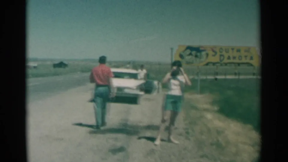 1959 8mm road-trip frame showing a woman with a camera near the South Dakota sign while a man moves toward the parked white car.