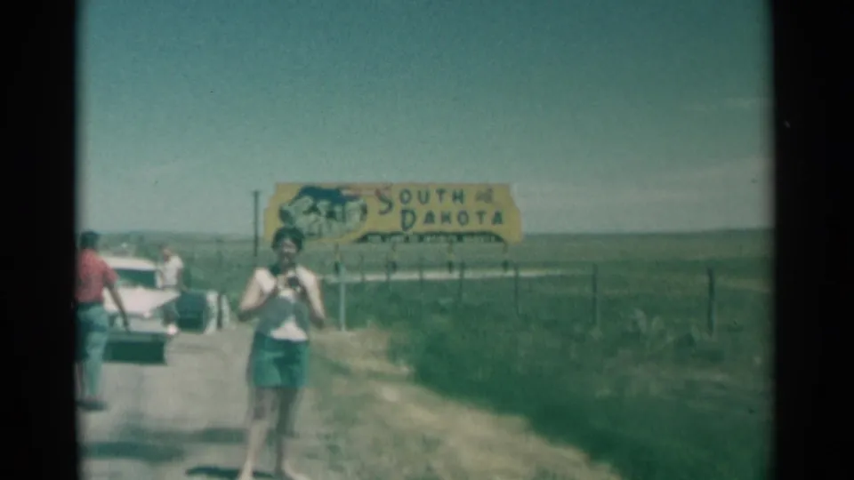South Dakota road-trip still from 1959 with a woman holding a camera in front of the yellow state sign and open prairie beyond.