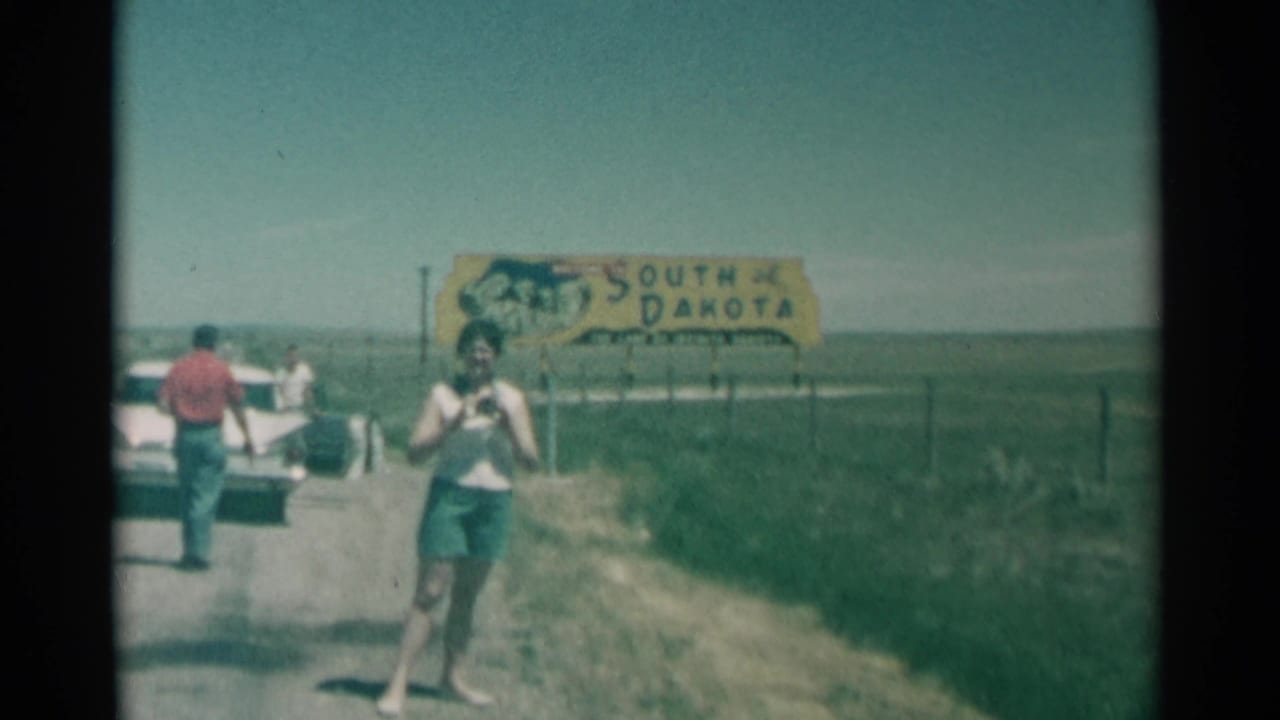 Woman holding a camera walks on a rural road near a South Dakota sign and parked white car.