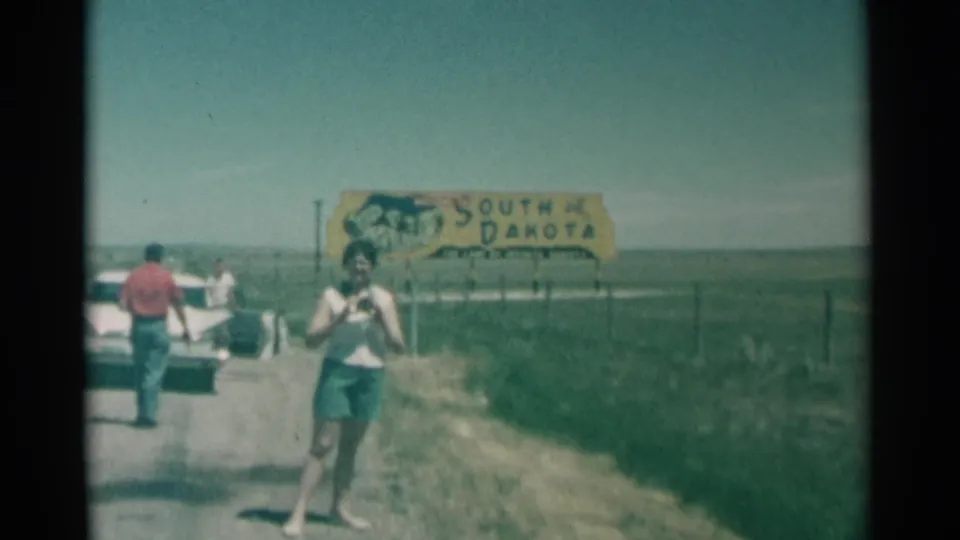 In 1959 South Dakota home-movie footage, a woman with a camera approaches the filmer beside a rural road, with a white car and state sign behind her.