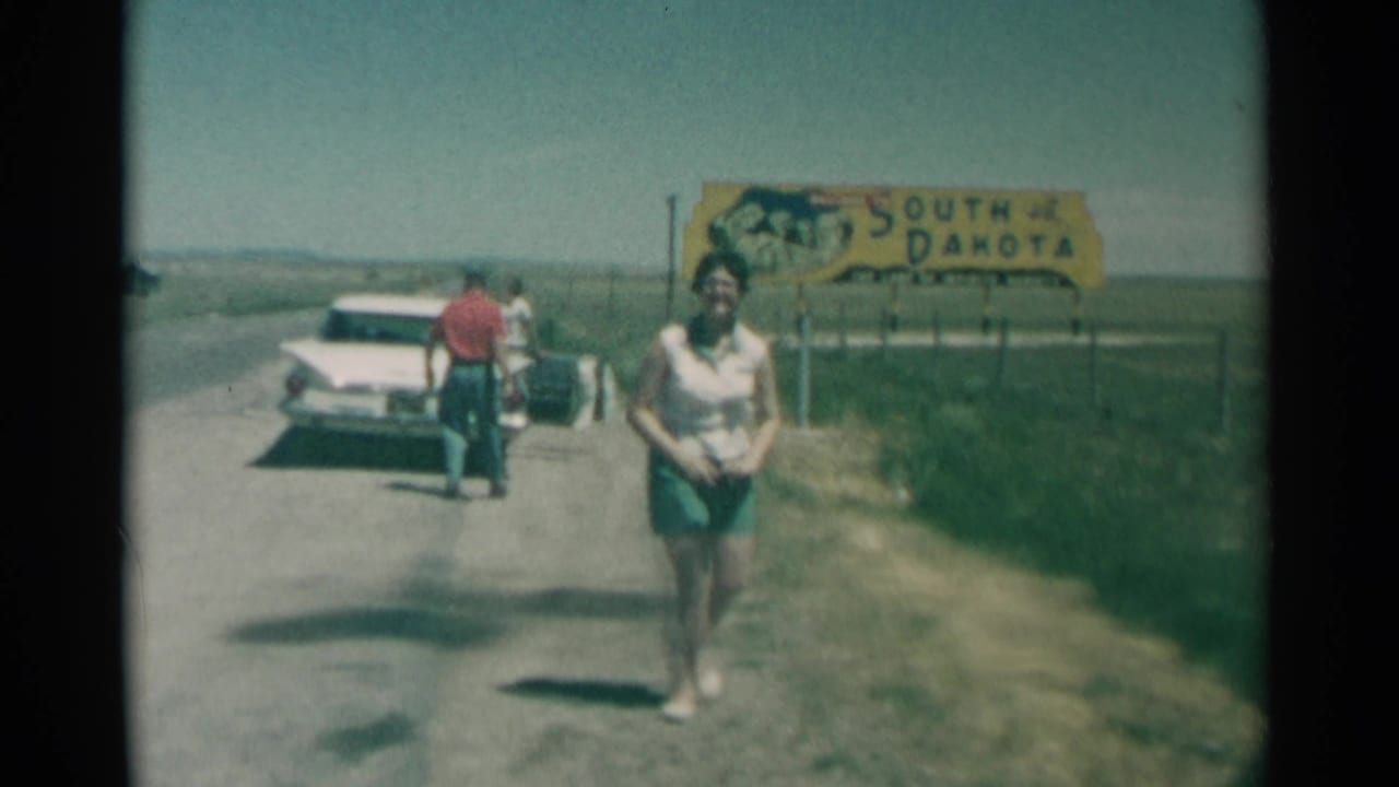 Woman stands closer to the camera as a man and another person remain near a white car and South Dakota sign.
