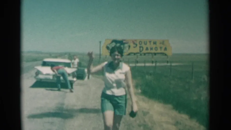 A woman waves during the South Dakota roadside stop while a man bends at the rear of the white car and the yellow state sign remains visible.