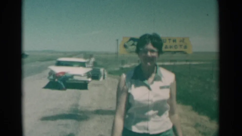 The woman walks close past the filmer in the 1959 South Dakota roadside scene, with the parked white car and yellow state sign in soft focus behind her.