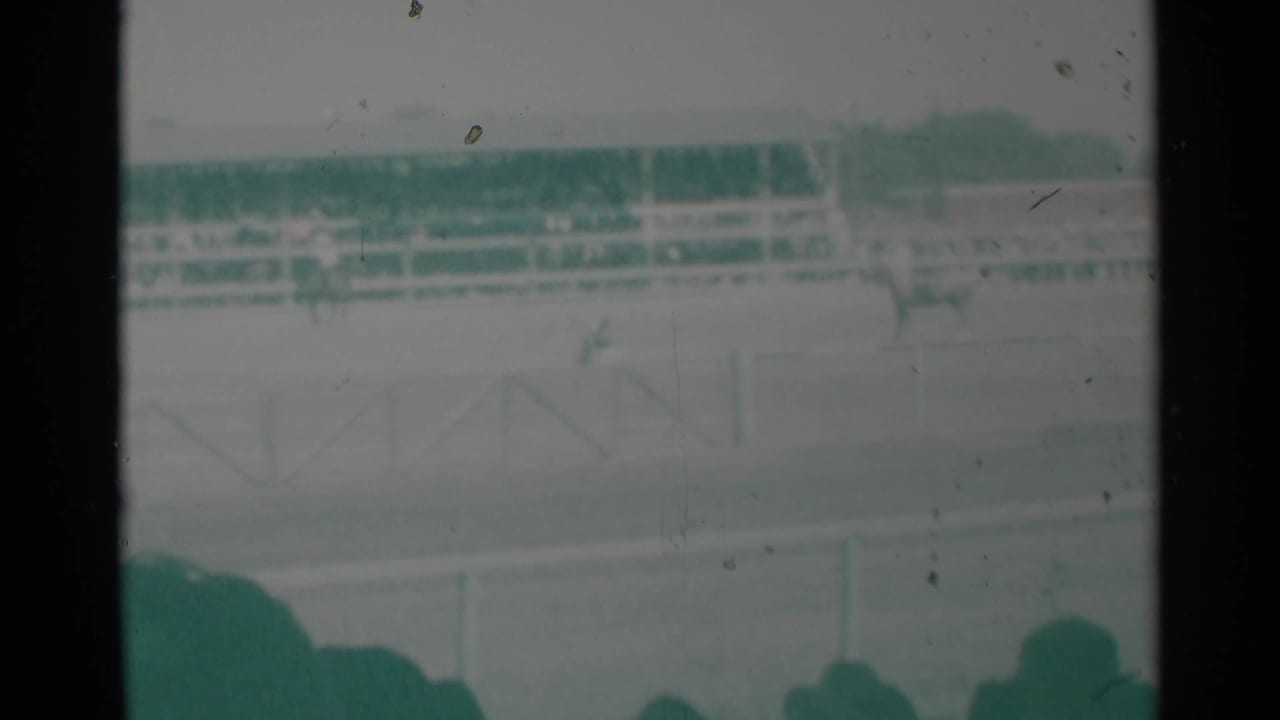 Spectators behind a rail watch a racetrack with a grandstand in the distance.