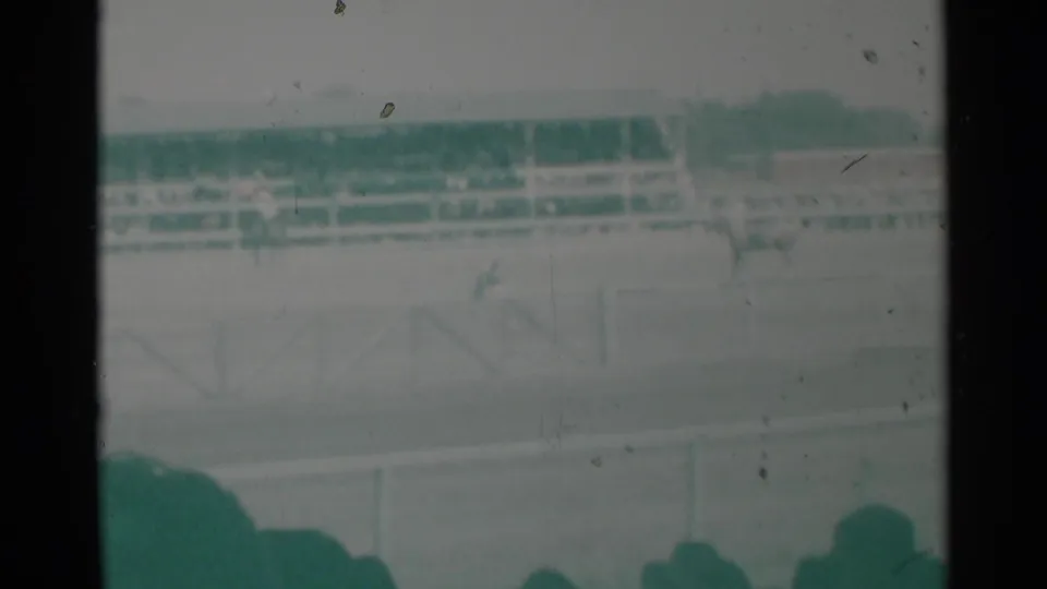 1960 Cheyenne racetrack view from behind spectators, with railings, a distant grandstand, and horses faintly visible on the field.